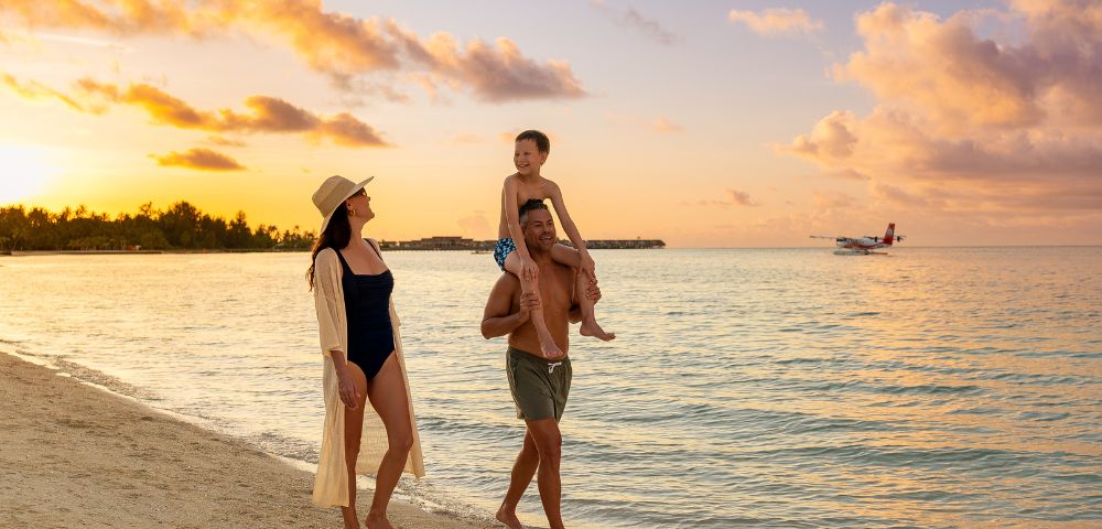A family walks along a beach at sunset, with warm, golden sky hues. The child sits on the father's shoulders, and a small plane flies over the sea.