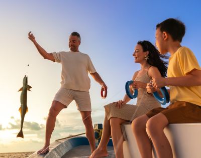 A family enjoying a joyful moment on a boat at sunset. A smiling man holds a fishing line with a large fish. A woman and a child watch with excitement.