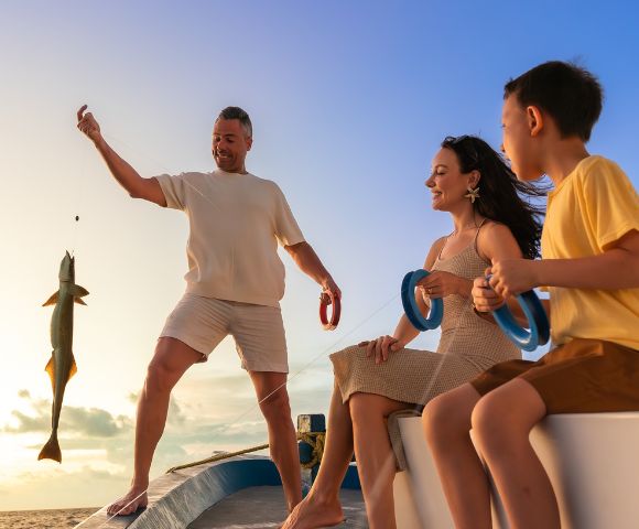 A family enjoying a joyful moment on a boat at sunset. A smiling man holds a fishing line with a large fish. A woman and a child watch with excitement.