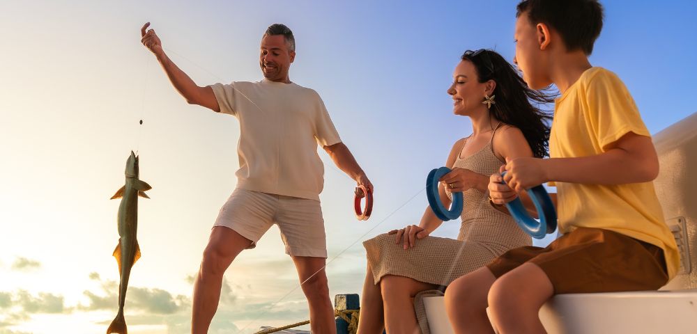 A family enjoying a joyful moment on a boat at sunset. A smiling man holds a fishing line with a large fish. A woman and a child watch with excitement.