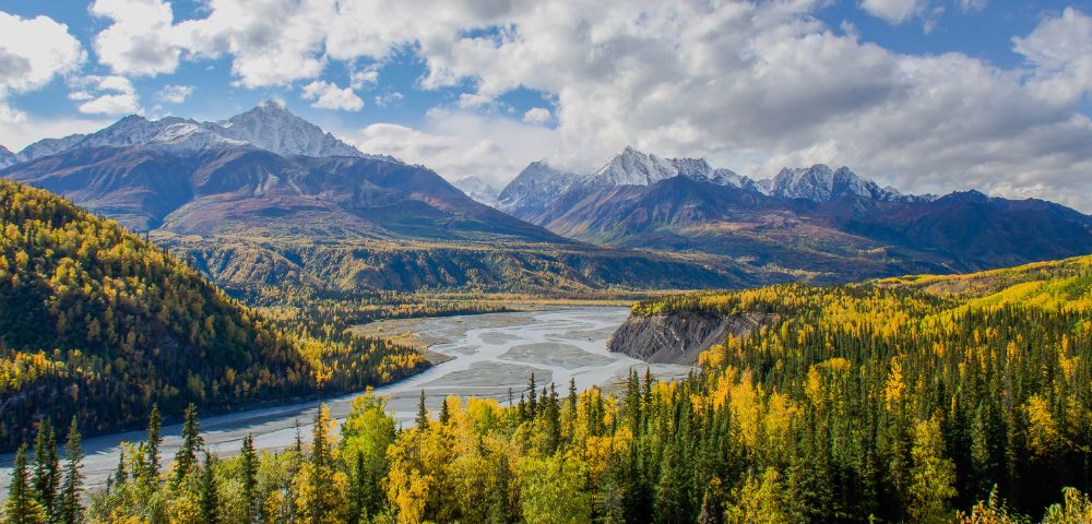 Panoramic view of a serene mountain landscape with a winding river, surrounded by autumnal trees. Snow-capped peaks under a partly cloudy sky evoke tranquility.
