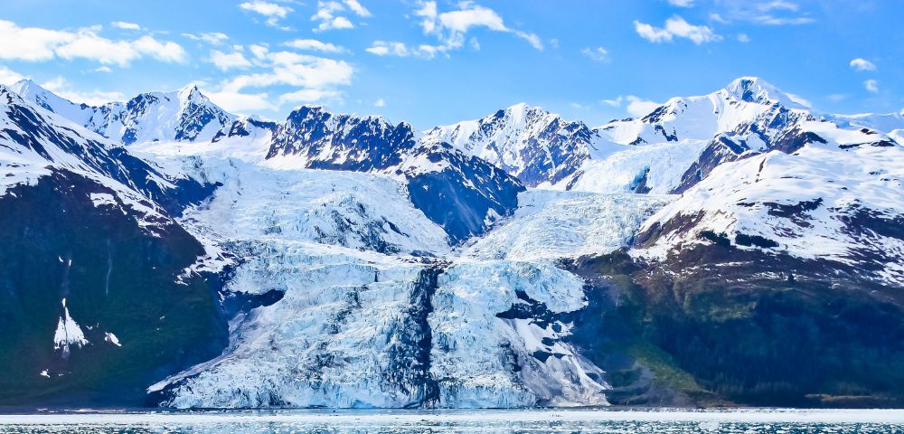 Snow-capped mountains with a central glacier descending into a serene icy body of water under a bright blue sky, conveying a sense of tranquility and grandeur.