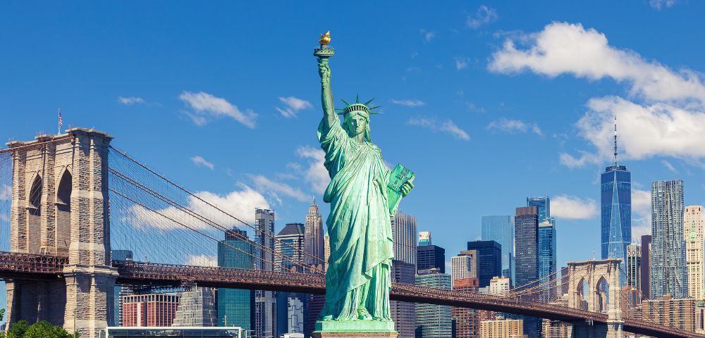 The image shows the Statue of Liberty in the foreground with the Brooklyn Bridge and New York City skyline under a clear blue sky in the background.