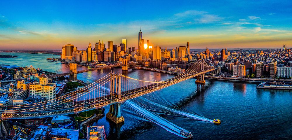 Aerial view of Manhattan at sunset, showcasing a vibrant skyline with glowing skyscrapers. The Brooklyn Bridge spans the East River, under a colorful sky.
