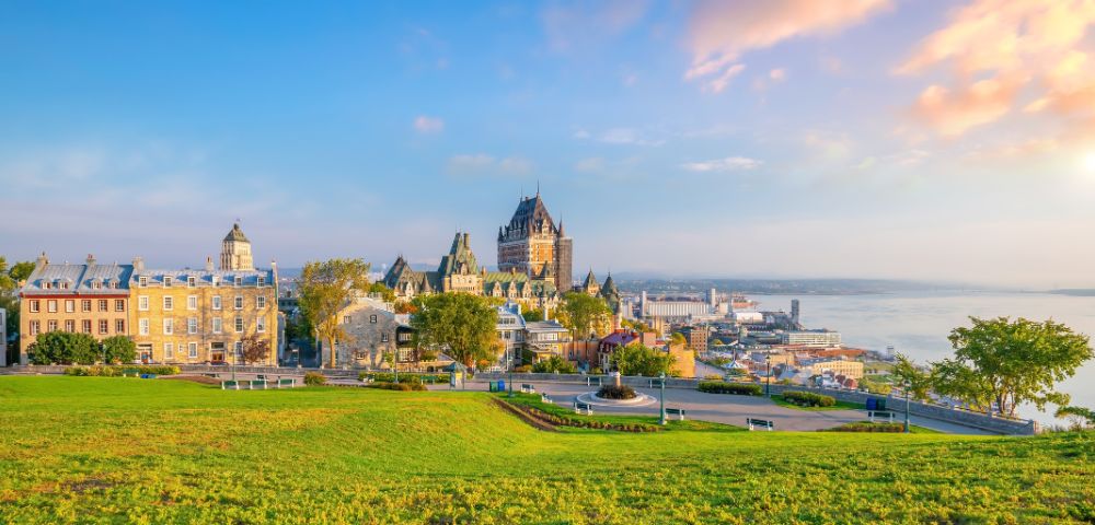 Panoramic view of Quebec City at sunrise, featuring historic buildings with the iconic Château Frontenac. The scene exudes tranquility and charm, with a lush green foreground and a calm river in the background.