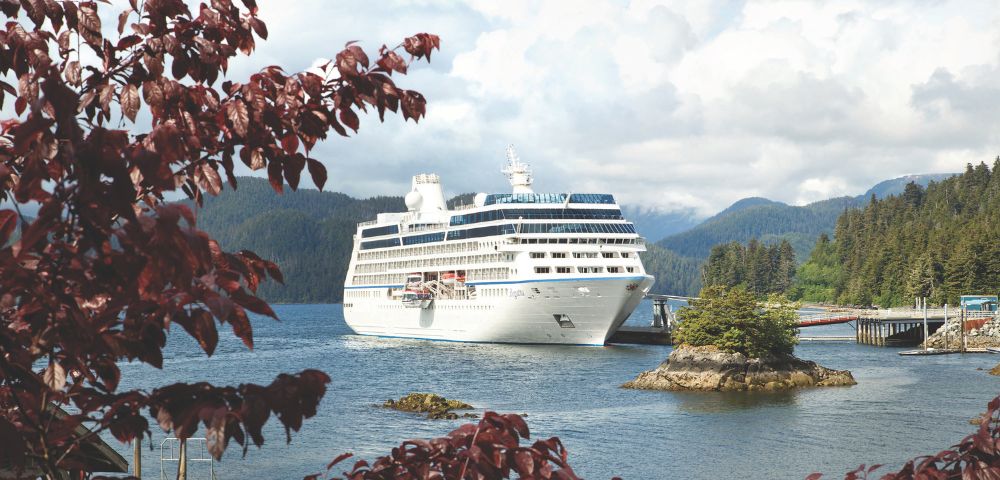 A large white cruise ship docked near a lush, forested shoreline under a cloudy sky. Foreground features red leaves framing the scene, creating a tranquil atmosphere.