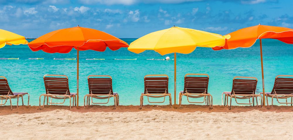 Five empty lounge chairs under vibrant orange and yellow umbrellas on a sandy beach, overlook a calm, turquoise sea under a clear blue sky.
