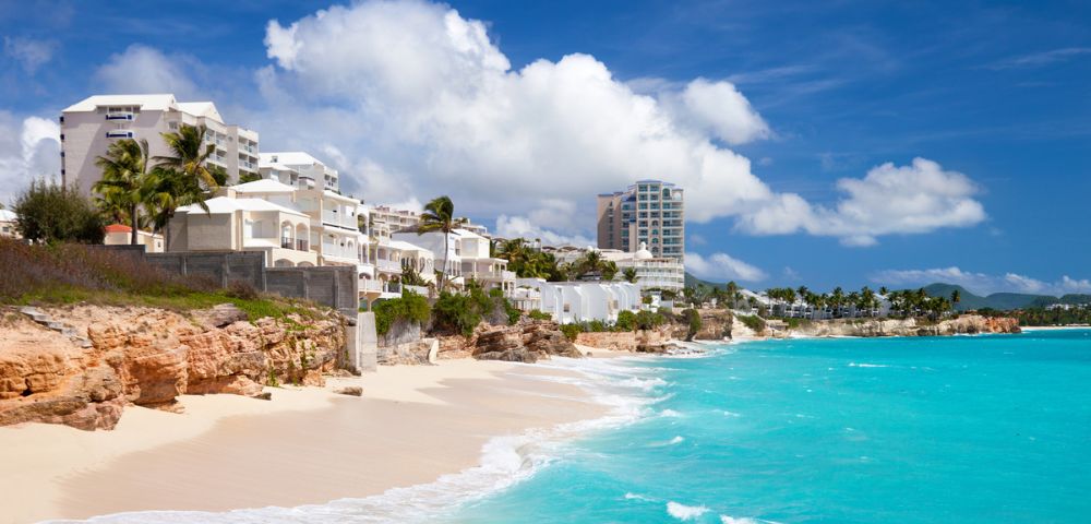 Coastal scene with turquoise water, white sandy beach, and cliffside buildings under a bright blue sky. The atmosphere is tranquil and inviting.