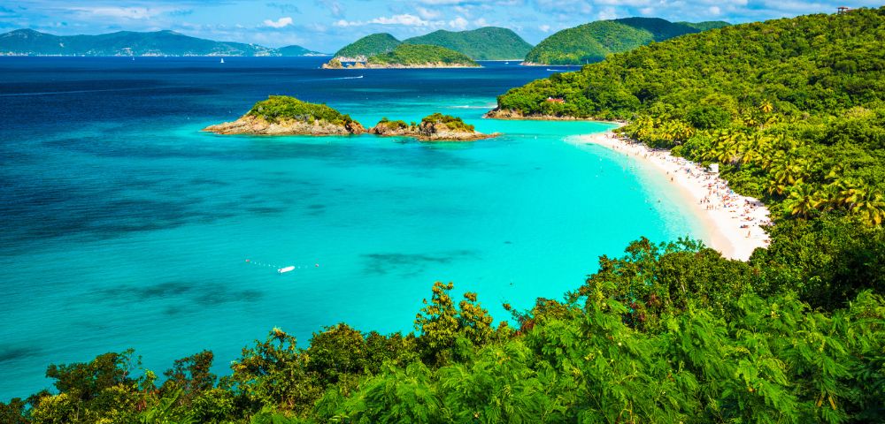 A stunning tropical beach with vibrant turquoise waters surrounded by lush green hills. The sky is bright blue with scattered fluffy clouds, conveying tranquility.