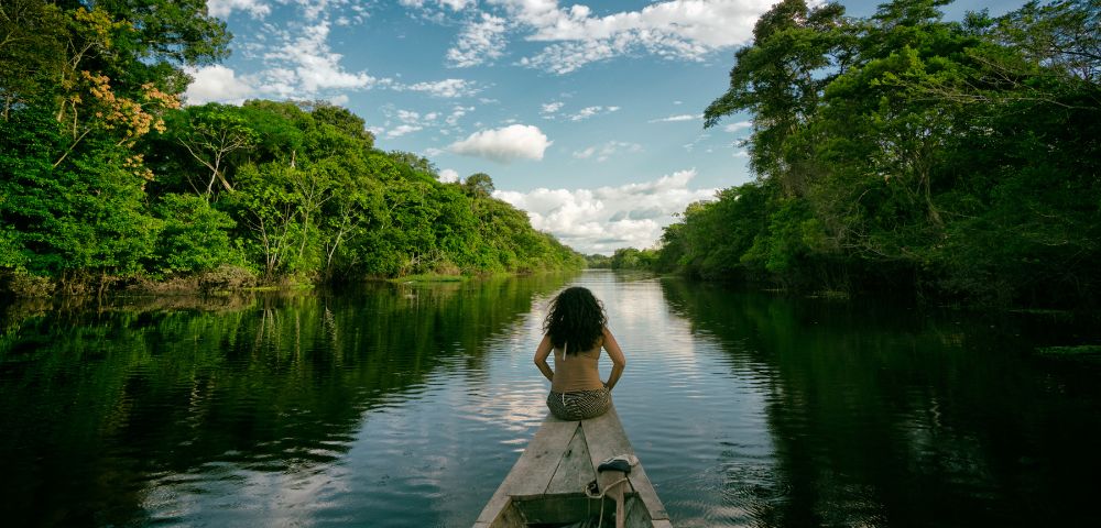 A woman sits on a boat gliding through a river surrounded by lush green forest under a partly cloudy sky, conveying tranquility and adventure.
