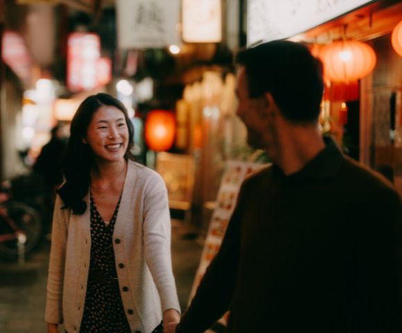 A couple walks hand in hand through a warmly lit street adorned with lanterns and signs. They smile at each other, conveying a joyful, relaxed atmosphere.