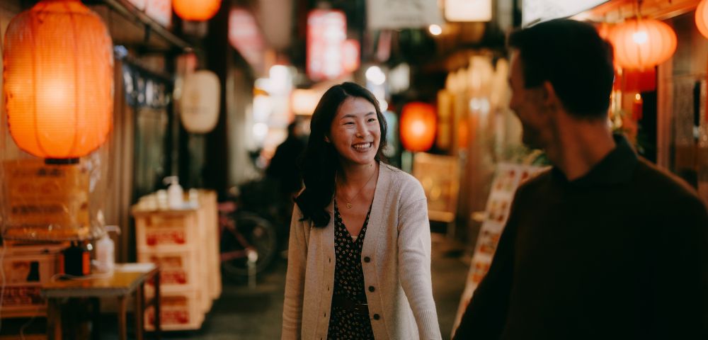 A couple walks hand in hand through a warmly lit street adorned with lanterns and signs. They smile at each other, conveying a joyful, relaxed atmosphere.