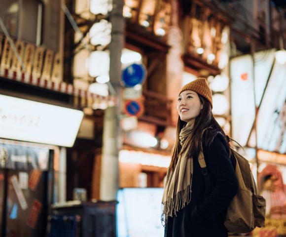 A woman in a beanie and scarf smiles while walking through a vibrant, illuminated urban street at night, exuding a sense of adventure and contentment.