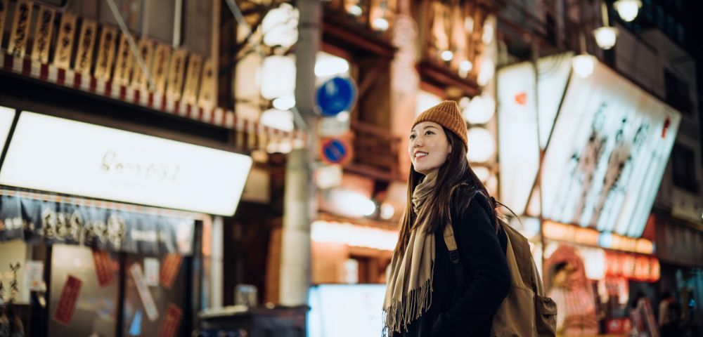 A woman in a beanie and scarf smiles while walking through a vibrant, illuminated urban street at night, exuding a sense of adventure and contentment.