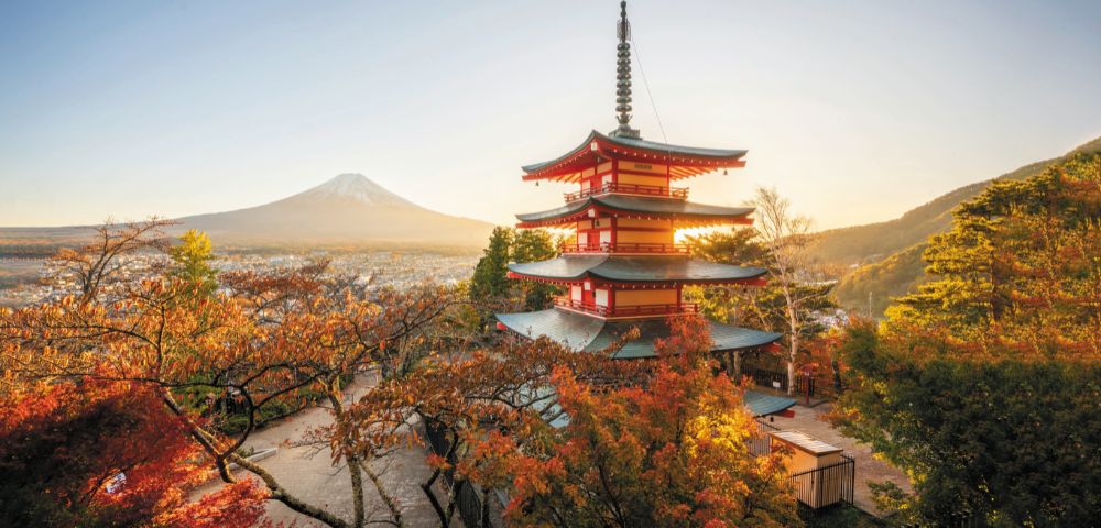 A serene landscape featuring a red pagoda surrounded by vibrant autumn trees. Mount Fuji is visible in the background under a clear, golden sky.