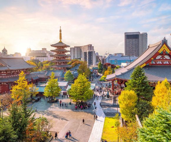 A serene aerial view of a Japanese temple complex with a pagoda, lush trees, and busy pathways. The skyline shows modern buildings under a warm sunset.