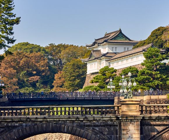 A historic Japanese palace with curved roofs stands behind a stone bridge surrounded by lush green trees under a clear blue sky, conveying tranquility.