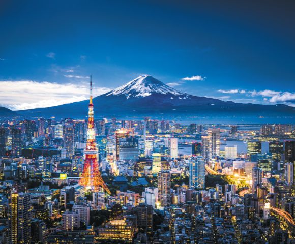 Tokyo skyline at dusk, with an illuminated Tokyo Tower in the foreground. Mount Fuji rises majestically in the background under a blue sky.