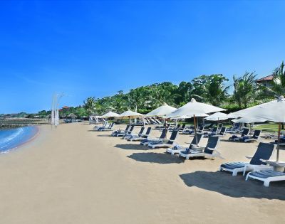 A serene beach scene with rows of lounge chairs and white umbrellas on golden sand. Calm blue ocean waves and a clear sky create a tranquil atmosphere.