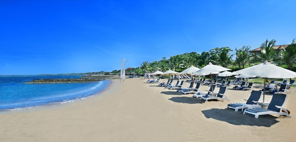 A serene beach scene with rows of lounge chairs and white umbrellas on golden sand. Calm blue ocean waves and a clear sky create a tranquil atmosphere.
