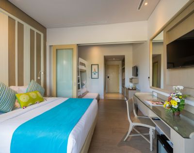 Modern hotel room with a double bed featuring a turquoise throw and decorative pillows. Bunk beds are visible through a sliding door. A desk with flowers and a TV adorn the space, exuding a cozy and inviting ambiance.