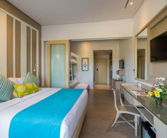 Modern hotel room with a double bed featuring a turquoise throw and decorative pillows. Bunk beds are visible through a sliding door. A desk with flowers and a TV adorn the space, exuding a cozy and inviting ambiance.