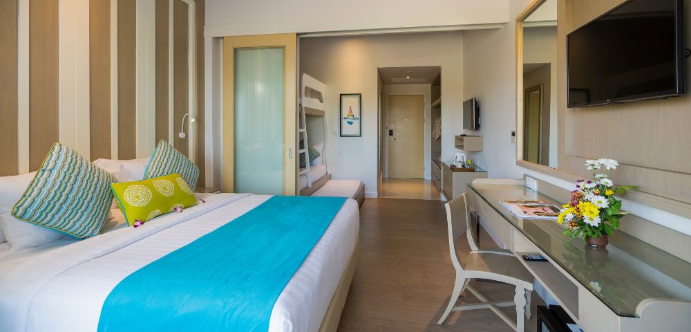 Modern hotel room with a double bed featuring a turquoise throw and decorative pillows. Bunk beds are visible through a sliding door. A desk with flowers and a TV adorn the space, exuding a cozy and inviting ambiance.