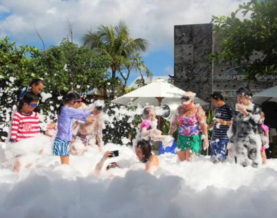A joyful group of people, including children and adults, are playing in abundant white foam outdoors. The scene is lively and fun, with greenery, umbrellas, and a rock wall in the background under a bright, sunny sky.