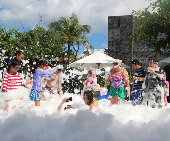 A joyful group of people, including children and adults, are playing in abundant white foam outdoors. The scene is lively and fun, with greenery, umbrellas, and a rock wall in the background under a bright, sunny sky.