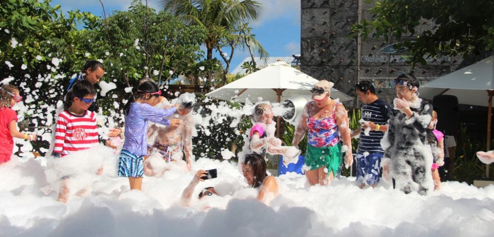 A joyful group of people, including children and adults, are playing in abundant white foam outdoors. The scene is lively and fun, with greenery, umbrellas, and a rock wall in the background under a bright, sunny sky.