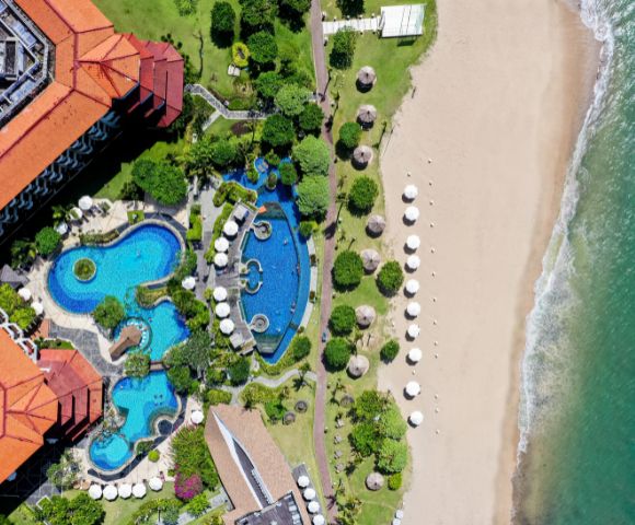 Aerial view of a beachfront resort with turquoise winding pools, lush greenery, and red-roofed buildings. White umbrellas line the sandy beach by the ocean.