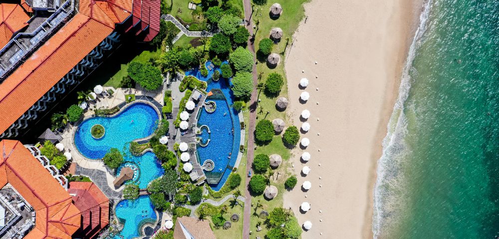 Aerial view of a beachfront resort with turquoise winding pools, lush greenery, and red-roofed buildings. White umbrellas line the sandy beach by the ocean.