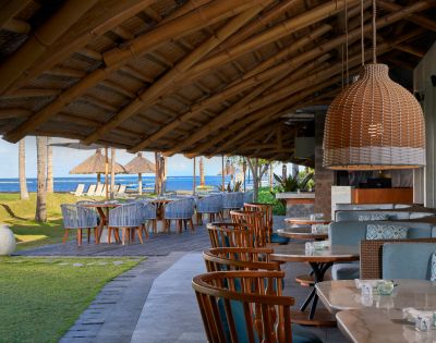 Outdoor restaurant with wood tables and chairs under a thatched roof, overlooking a beach with straw umbrellas and blue sea. Relaxing and inviting atmosphere.