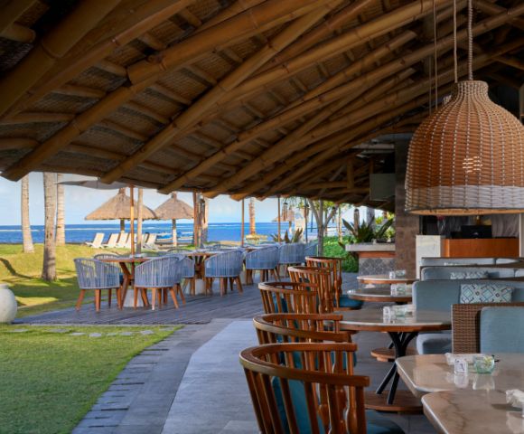 Outdoor restaurant with wood tables and chairs under a thatched roof, overlooking a beach with straw umbrellas and blue sea. Relaxing and inviting atmosphere.