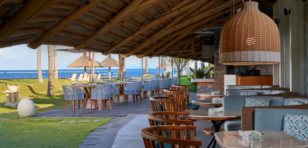 Outdoor restaurant with wood tables and chairs under a thatched roof, overlooking a beach with straw umbrellas and blue sea. Relaxing and inviting atmosphere.