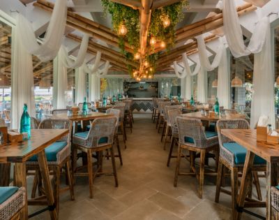 Spacious open-air restaurant with wicker chairs, wooden tables, and teal cushions. Hanging lights and greenery create a cozy, inviting atmosphere.
