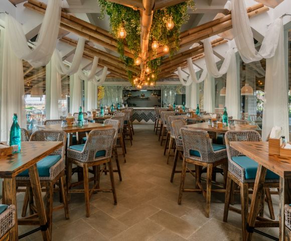 Spacious open-air restaurant with wicker chairs, wooden tables, and teal cushions. Hanging lights and greenery create a cozy, inviting atmosphere.