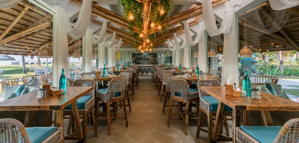 Spacious open-air restaurant with wicker chairs, wooden tables, and teal cushions. Hanging lights and greenery create a cozy, inviting atmosphere.