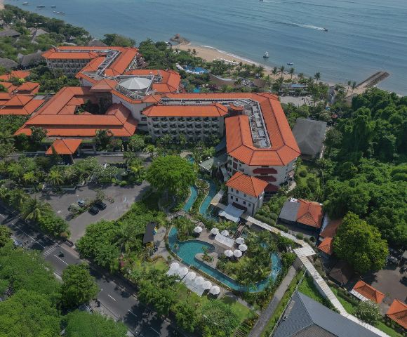 Aerial view of a coastal resort with terracotta-roofed buildings surrounded by lush greenery. A winding pool and beach are visible, evoking a tropical vibe.