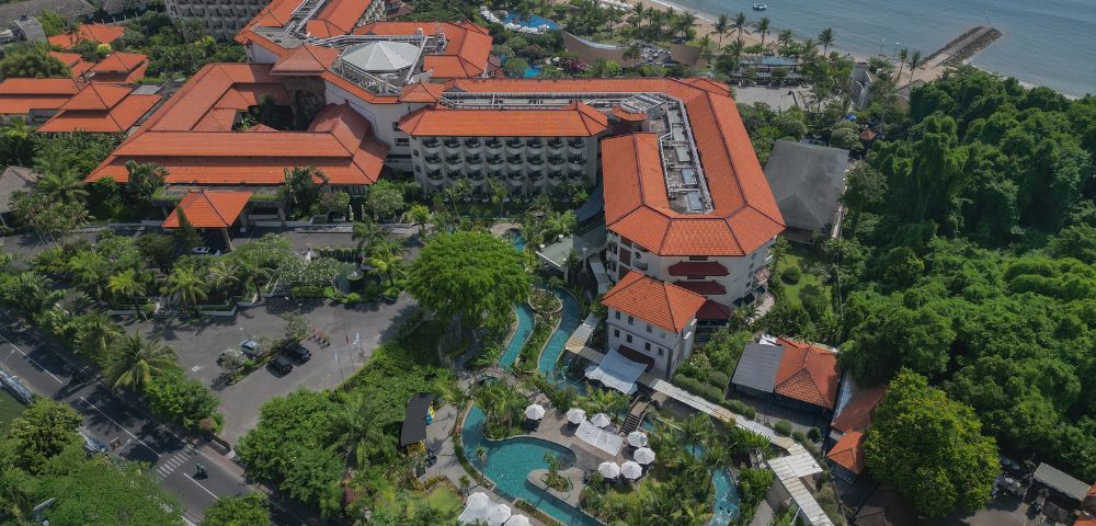 Aerial view of a coastal resort with terracotta-roofed buildings surrounded by lush greenery. A winding pool and beach are visible, evoking a tropical vibe.