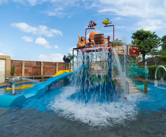 Children's water park with vibrant slides and cascading water. An octopus sculpture sits atop. The scene is playful, under a bright blue sky.