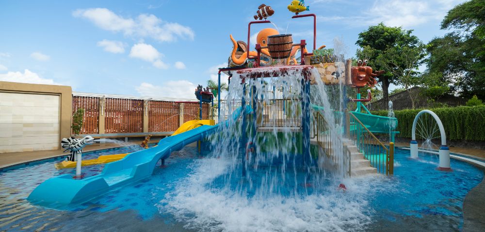 Children's water park with vibrant slides and cascading water. An octopus sculpture sits atop. The scene is playful, under a bright blue sky.