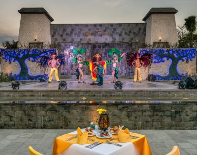 A vibrant outdoor performaance features five dancers in colorful costumes with feathered headpieces on a stage, viewed from a dining table setup.