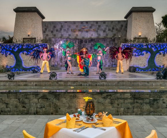 A vibrant outdoor performance features five dancers in colorful costumes with feathered headpieces on a stage, viewed from a dining table setup.