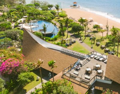 Aerial view of a beachfront resort with lush greenery, a pool complex, and a sandy beach. There's a spacious terrace with lounge chairs, evoking relaxation.