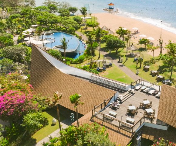 Aerial view of a beachfront resort with lush greenery, a pool complex, and a sandy beach. There's a spacious terrace with lounge chairs, evoking relaxation.