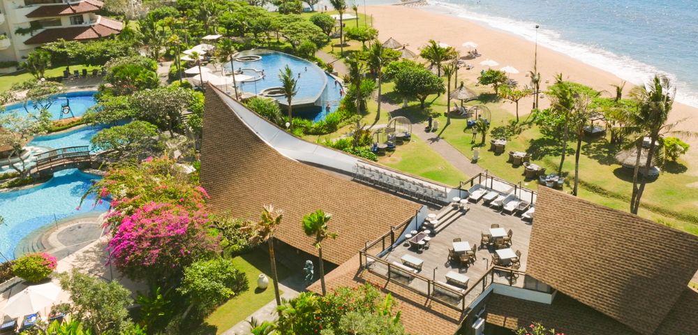 Aerial view of a beachfront resort with lush greenery, a pool complex, and a sandy beach. There's a spacious terrace with lounge chairs, evoking relaxation.