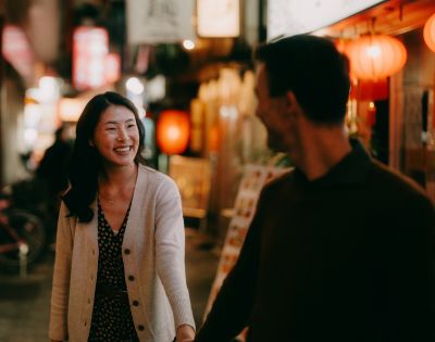 A woman and man smile at each other, holding hands, as they walk through a warmly lit street with lanterns, creating a joyful and romantic atmosphere.
