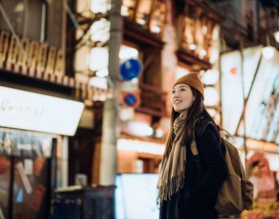 A woman in a beanie and scarf smiles while walking in a vibrant, illuminated urban street at night, conveying warmth and exploration.
