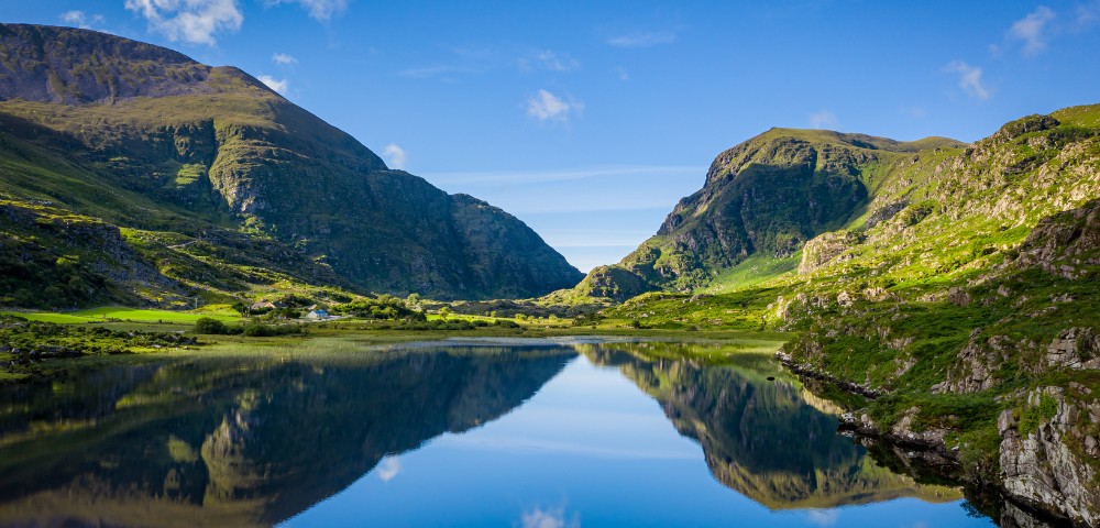 A serene lake reflects lush green mountains under a clear blue sky, creating a tranquil and picturesque landscape with vibrant colors and peaceful ambiance.
